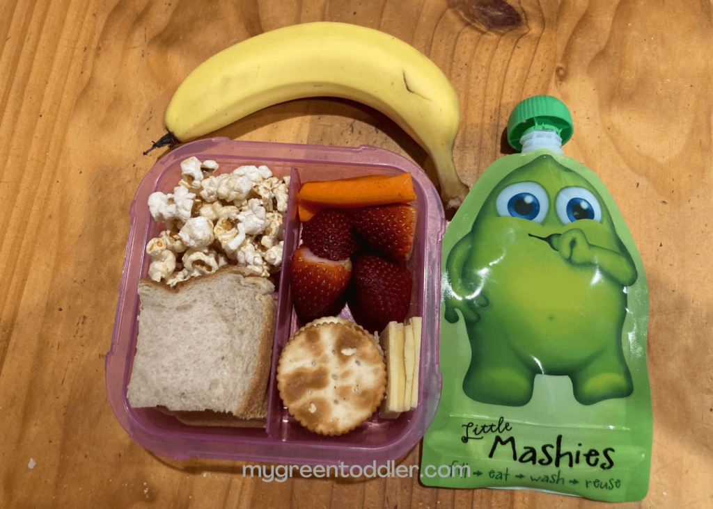 Lunchbox packed with sandwich, popcorn, carrot sticks, strawberries, cheese and crackers alongside a reusable yoghurt pouch and banana.