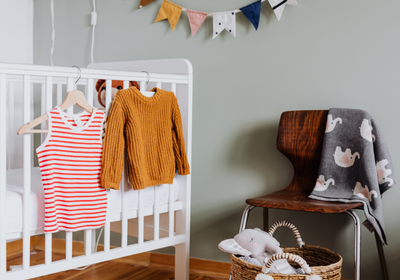 Second hand baby clothes. A nursery with a stripey vest and woolen jumper on coat hangers hanging on the side of a cot next to a chair with a throw rug and basket of soft toys.